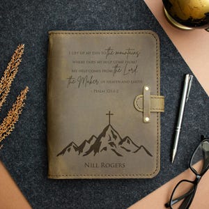 Könnte beinhalten: Ein braunes Lederjournal mit einem Berg- und Kreuzdesign sowie dem Text "I lift up my eyes to the mountains..." und "Nill Rogers". Das Journal hat einen Riemenverschluss und liegt auf einer dunklen Oberfläche.