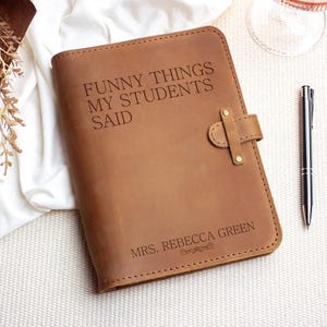 Op de afbeelding: Een bruin leren notitieboek met de tekst "FUNNY THINGS MY STUDENTS SAID" en "MRS. REBECCA GREEN" gegraveerd op de kaft. Het notitieboek heeft een riemsluiting en ligt naast een zilveren pen.