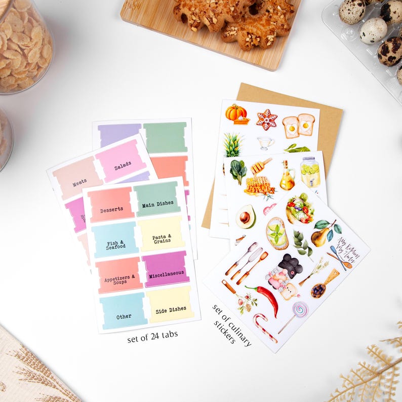 K&ouml;nnte beinhalten: Ein Set aus 24 farbigen K&uuml;chenetiketten mit Kategorien wie "Fleisch", "Salate" und "Desserts". Au&szlig;erdem ein Set kulinarischer Aufkleber mit Lebensmittelillustrationen, darunter ein K&uuml;rbis, eine Avocado und Honig. Die Etiketten und Aufkleber liegen auf einer wei&szlig;en Oberfl&auml;che.
