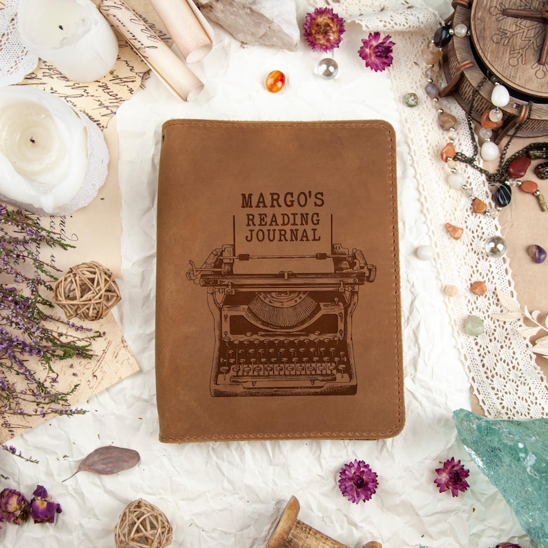 Puede incluir: Un diario de cuero marr&oacute;n con el texto "MARGO'S READING JOURNAL" y una ilustraci&oacute;n de m&aacute;quina de escribir. El diario es rectangular y est&aacute; cerrado. El fondo incluye elementos decorativos como velas, flores y papel vintage.