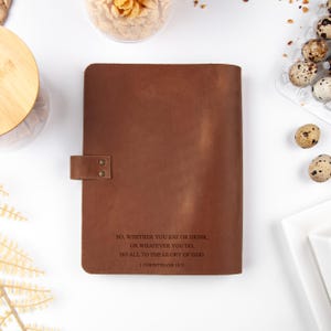 K&ouml;nnte beinhalten: Ein braunes Lederjournal mit einem Schnappverschluss. Das Journal tr&auml;gt den Text "SO, WHETHER YOU EAT OR DRINK, OR WHATEVER YOU DO, DO ALL TO THE GLORY OF GOD. I CORINTHIANS 10:11". Das Journal liegt auf einer wei&szlig;en Oberfl&auml;che.