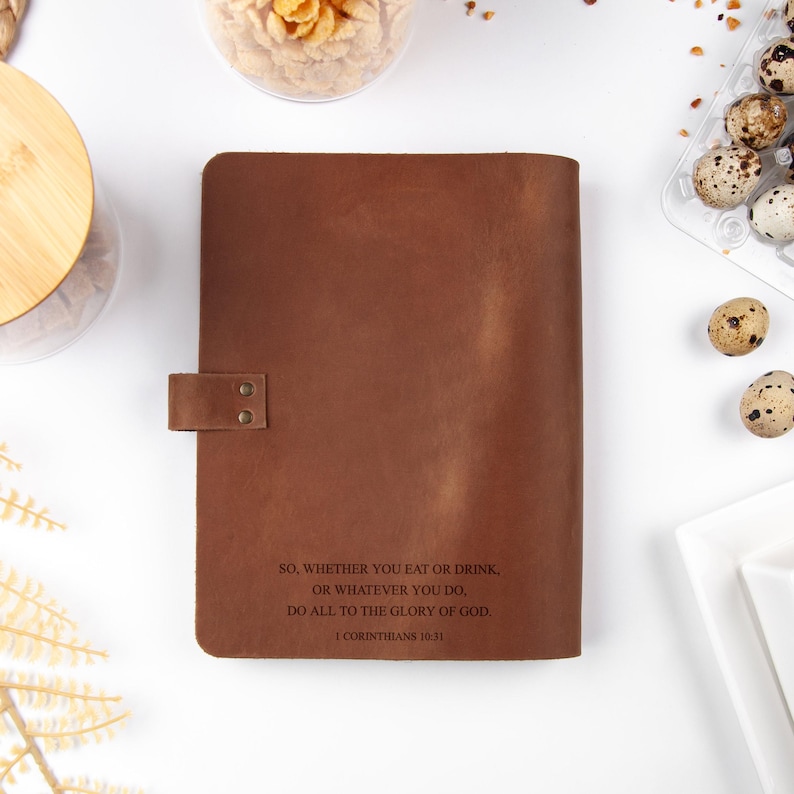 K&ouml;nnte beinhalten: Ein braunes Lederjournal mit einem Schnappverschluss. Das Journal tr&auml;gt den Text "SO, WHETHER YOU EAT OR DRINK, OR WHATEVER YOU DO, DO ALL TO THE GLORY OF GOD. I CORINTHIANS 10:11". Das Journal liegt auf einer wei&szlig;en Oberfl&auml;che.