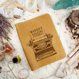 Puede incluir: Un diario de cuero color canela con el texto "MARGO'S READING JOURNAL" y una ilustraci&oacute;n de m&aacute;quina de escribir. El diario est&aacute; rodeado de elementos decorativos como velas, flores secas y encaje vintage.