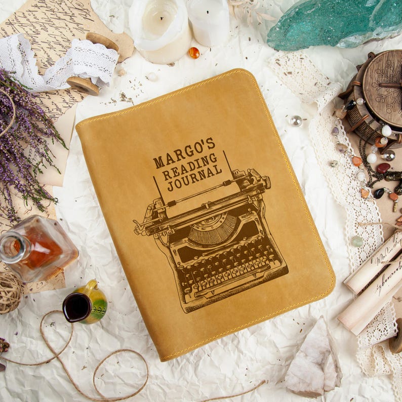 Puede incluir: Un diario de cuero color canela con el texto "MARGO'S READING JOURNAL" y una ilustraci&oacute;n de m&aacute;quina de escribir. El diario est&aacute; rodeado de elementos decorativos como velas, flores secas y encaje vintage.