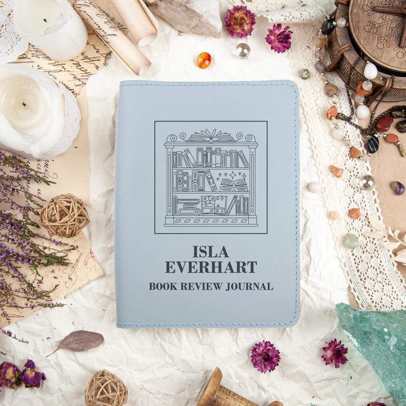 Puede incluir: Un diario de rese&ntilde;as de libros azul claro con el texto "ISLA EVERHART BOOK REVIEW JOURNAL". La portada presenta una ilustraci&oacute;n en blanco y negro de una estanter&iacute;a. El diario est&aacute; rodeado de objetos decorativos.