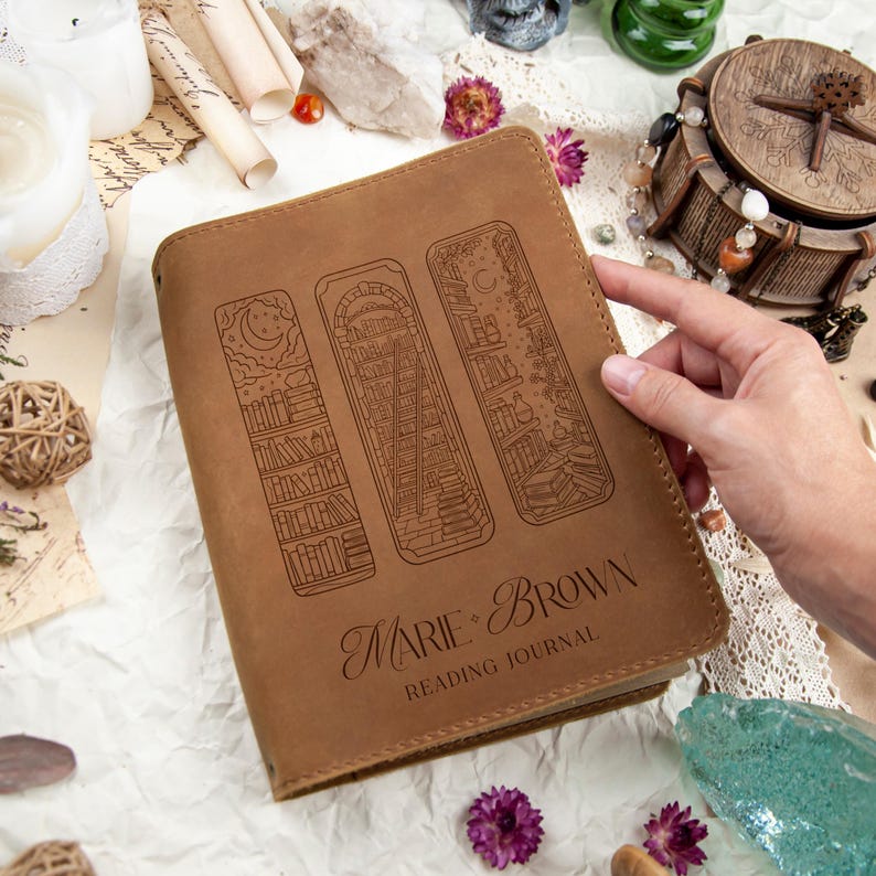 Puede incluir: Un diario de lectura de cuero marr&oacute;n con tres dise&ntilde;os de marcap&aacute;ginas grabados. La portada del diario presenta el texto "Marie + Brown READING JOURNAL". Los dise&ntilde;os de marcap&aacute;ginas incluyen una luna y estrellas, una biblioteca con una escalera y un cielo nocturno con libros.
