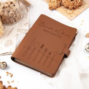 K&ouml;nnte beinhalten: Ein braunes Leder-Rezeptbuch mit dem Text "Brenda & Oliver Recipe Collection" und Besteckillustrationen. Das Buch hat einen Riemenverschluss und ist von Zutaten wie Zuckerw&uuml;rfeln und Keksen umgeben.