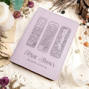 Puede incluir: Un diario de lectura lila con tres dise&ntilde;os de marcap&aacute;ginas ilustrados. La portada del diario presenta el texto "Marie & Brown Reading Journal" en una elegante escritura. Las ilustraciones representan estanter&iacute;as con elementos celestiales.