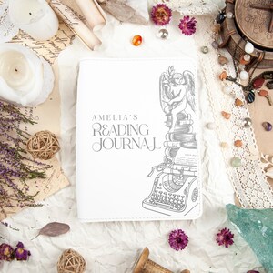 Puede incluir: Un diario de lectura blanco con el texto "AMELIA'S READING JOURNAL" en gris. La portada presenta una ilustraci&oacute;n gris de un &aacute;ngel, libros y una m&aacute;quina de escribir. El diario est&aacute; rodeado de objetos decorativos.