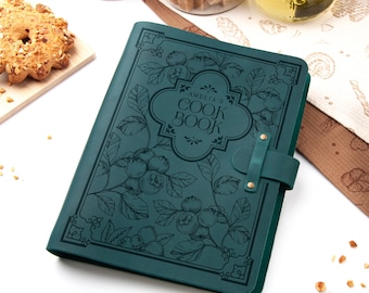 Könnte beinhalten: Ein türkisfarbenes Kochbuch mit dem Text "AMELIA'S COOK BOOK" in einem dekorativen Rahmen. Das Cover zeigt ein botanisches Design aus Blaubeeren und Blättern. Ein Lederriemen mit Messingnieten sichert das Buch. Das Kochbuch liegt auf einer weißen Oberfläche.