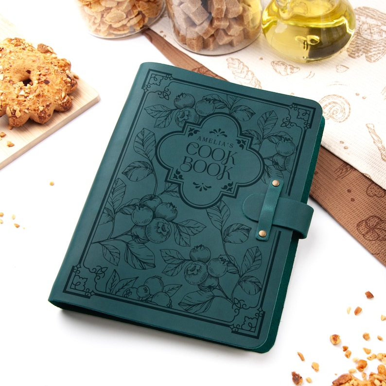 K&ouml;nnte beinhalten: Ein t&uuml;rkisfarbenes Kochbuch mit dem Text "AMELIA'S COOK BOOK" in einem dekorativen Rahmen. Das Cover zeigt ein botanisches Design aus Blaubeeren und Bl&auml;ttern. Ein Lederriemen mit Messingnieten sichert das Buch. Das Kochbuch liegt auf einer wei&szlig;en Oberfl&auml;che.