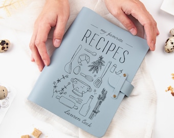 May include: A light blue recipe book with the words "my favorite RECIPES" and illustrated kitchen tools. The book has a strap closure and the name "Lauren Cook" is printed at the bottom. The book is held by two hands.