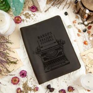Puede incluir: Un diario de cuero marr&oacute;n con el texto "MARGO'S READING JOURNAL" y una ilustraci&oacute;n de m&aacute;quina de escribir. El diario est&aacute; rodeado de flores secas, cristales y objetos decorativos, creando una est&eacute;tica vintage.