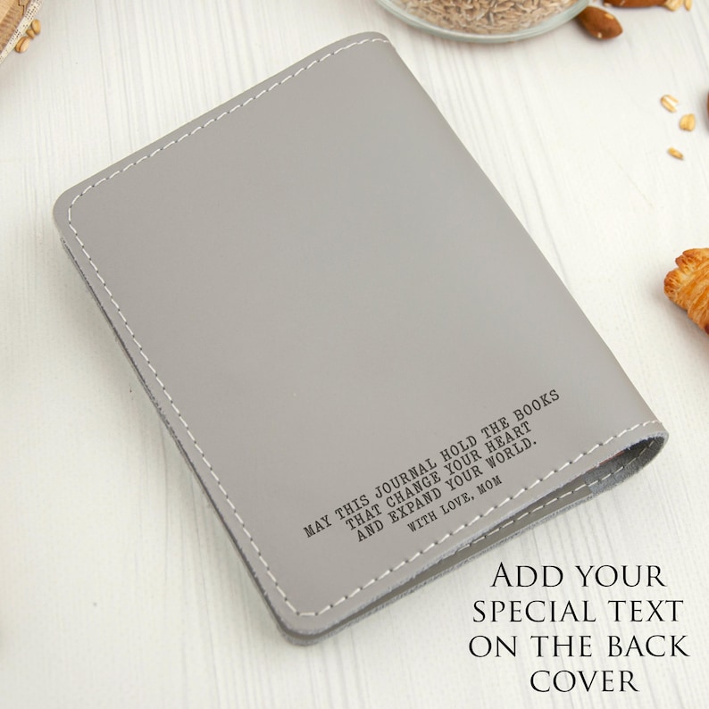 Puede incluir: Un diario gris con costuras blancas alrededor de los bordes. El diario tiene el texto "MAY THIS JOURNAL HOLD THE BOOKS THAT CHANGE YOUR HEART AND EXPAND YOUR WORLD. WITH LOVE, MOM." Texto adicional dice "ADD YOUR SPECIAL TEXT ON THE BACK COVER."