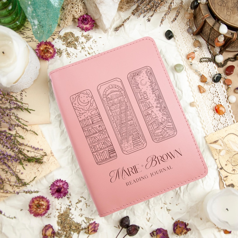 Puede incluir: Un diario de lectura rosa con tres dise&ntilde;os de marcap&aacute;ginas ilustrados. La portada del diario presenta el texto "Marie Brown Reading Journal". Las ilustraciones de los marcap&aacute;ginas representan estanter&iacute;as y elementos celestiales.