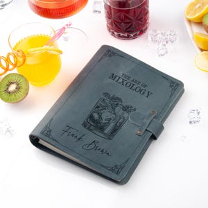 May include: A teal journal titled "THE ART OF MIXOLOGY" with the name "Frank Brown" is shown. The journal has a cocktail illustration and a strap closure. Drinks and garnishes are in the background, including a kiwi fruit.