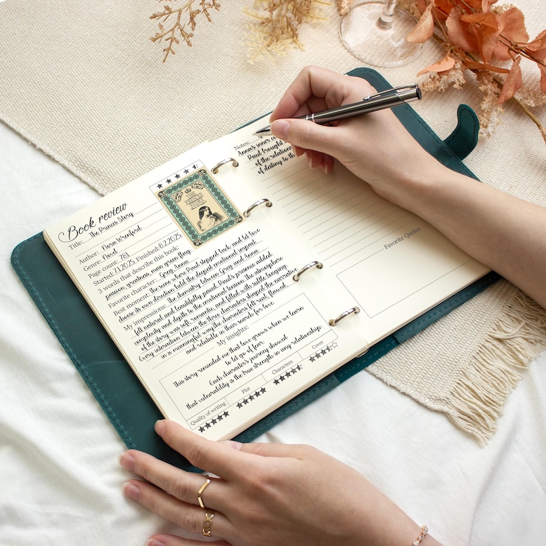 Puede incluir: Un diario de revisi&oacute;n de libros encuadernado en cuero color verde azulado est&aacute; abierto, con la mano de una persona sosteniendo un bol&iacute;grafo sobre las p&aacute;ginas rayadas. El diario tiene una tarjeta de revisi&oacute;n de libros con el t&iacute;tulo "The Prince Story".