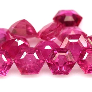 May include: A collection of faceted, hexagonal-shaped, deep pink gemstones. The gems are arranged closely together, with their multiple facets reflecting light. The gemstones are a vibrant shade of pink, and are set against a white background.
