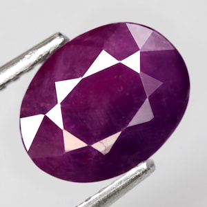 Op de afbeelding: Een gefacetteerde, ovaalvormige robijn edelsteen. De dieprode-paarse edelsteen wordt vastgehouden door zilveren pinnen, die de gepolijste oppervlakken en interne lichtreflecties laten zien. De slijping van de edelsteen onthult een reeks driehoekige facetten.