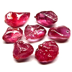 May include: Seven rough ruby gemstones in varying shapes and sizes. The stones are a deep red colour with a translucent quality. The gems are displayed against a white background, highlighting their natural textures. These gemstones are ideal for jewelry making.