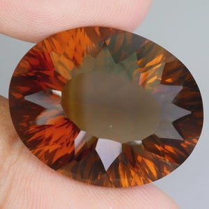 May include: A large, faceted gemstone with an oval shape. The gem features a blend of orange and brown hues, with a central area of lighter color. The facets create a starburst effect, reflecting light and adding depth to the stone.
