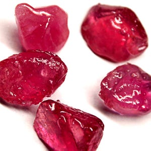 May include: Five raw, uncut ruby gemstones. The rubies are a deep, saturated red colour with a translucent quality. Each stone has a unique, irregular shape and a slightly glossy surface, suggesting they are freshly cut.