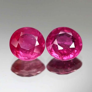 May include: Two round, faceted ruby gemstones. The rubies are a vibrant, deep pink-red colour, with multiple facets catching the light. The gemstones are displayed on a reflective surface, creating a mirrored effect.