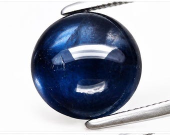 May include: A round, cabochon-cut deep blue gemstone with a smooth, polished surface and visible internal inclusions. The stone is held by a pair of silver-toned metal tweezers, showcasing its dark, saturated color and reflective, glossy finish.