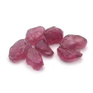 May include: A collection of seven small, raw, unpolished ruby gemstones. Each stone features an irregular, organic shape with a deep, saturated magenta-red color and a textured, matte surface appearance scattered across a plain white background.