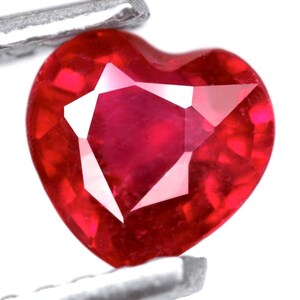 Może przedstawiać: Fasetowany rubin w kształcie serca. Rubin ma głęboki czerwony kolor i jest trzymany przez srebrne metalowe narzędzie. Kamień szlachetny ma wypolerowaną, odblaskową powierzchnię na białym tle.
