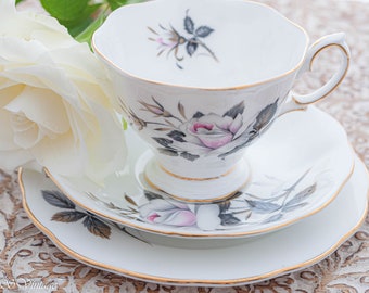 May include: A white teacup and saucer set with a floral design featuring pink and gray roses. The set has a gold trim around the edges.
