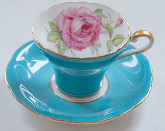May include: A teal teacup and saucer set with gold trim. The teacup interior features a pink rose design with green leaves. The set is likely made of porcelain and is suitable for serving tea. The saucer has a scalloped edge.
