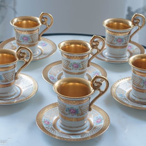 Może przedstawiać: Zestaw sześciu białych porcelanowych filiżanek do herbaty i spodków z pozłacanymi obwódkami i wzorami kwiatowymi. Każda filiżanka ma złote wnętrze i delikatny wzór kwiatowy na zewnątrz. Spodeczki mają pasujący wzór kwiatowy i pozłacane obwódki.