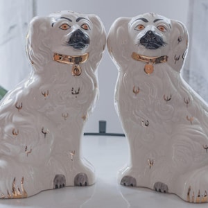 Peut inclure: Deux figurines de chiens en céramique blanche avec des détails dorés. Chaque chien a une moustache noire, des yeux orange et un collier doré. Les figurines sont assises, et les détails sont mis en valeur avec de la peinture dorée.
