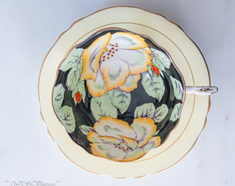 May include: A vintage teacup and saucer set with a black background and a floral design featuring yellow and white flowers with green leaves. The saucer is a pale yellow color with a gold trim.