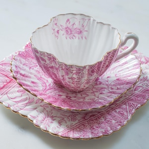May include: A vintage tea set comprising a teacup, saucer, and side plate. The white porcelain is decorated with a pink floral design and gold edging. The teacup has a scalloped rim and a curved handle. The set is on a white surface.