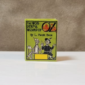 Op de afbeelding: Een kleine, rechthoekige doos met de titel "The Wonderful Wizard of Oz" van L. Frank Baum. De doos heeft een groene achtergrond met illustraties van personages uit het verhaal. Het woord "OZ" is rood en oranje.