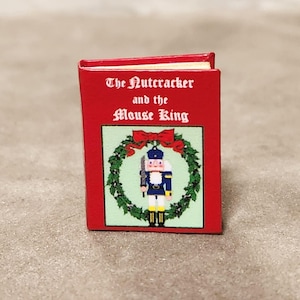 Pode incluir: Um livro em miniatura intitulado The Nutcracker and the Mouse King com uma capa vermelha brilhante. A frente apresenta uma ilustração central de um soldado quebra-nozes dentro de uma guirlanda verde com um laço vermelho, em texto branco.