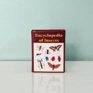 May include: A miniature book titled "Encyclopedia of Insects" with a maroon cover. The book's cover features illustrations of various insects, including a butterfly, ant, and ladybug. The background is a light teal color.