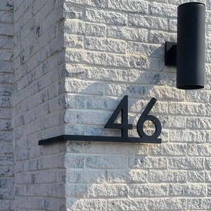 May include: Black metal house number plaque with the number 46 attached to a white brick wall.