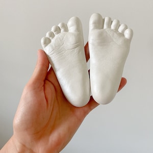 May include: Two white plaster baby foot prints, one in each hand, with the toes facing each other.