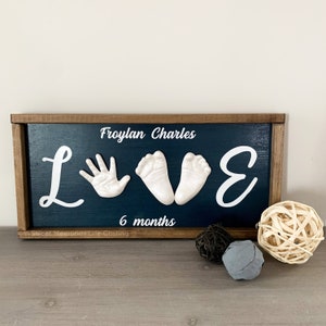 May include: A wooden sign with a dark blue background and white lettering. The sign says "LOVE" with a handprint and two footprints in the middle. The text "Froylan Charles" and "6 months" are also on the sign.