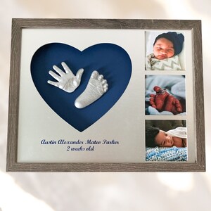 Pode incluir: Uma impressão de mão e pé prateadas em uma moldura em forma de coração azul. A moldura tem três fotos de um bebê. O texto abaixo do coração diz "Austin Alexander Mateo Parker 2 semanas".