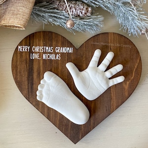 May include: A heart-shaped wooden plaque with a white handprint and footprint, a white inscription that reads "Merry Christmas Grandma! Love, Nicholas" and the text "Sweet Memories Life Casting" in the top right corner.
