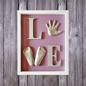 May include: A white frame with a pink background and gold-colored letters spelling "LOVE". The "O" is made up of two baby footprints and the "V" is a baby handprint.