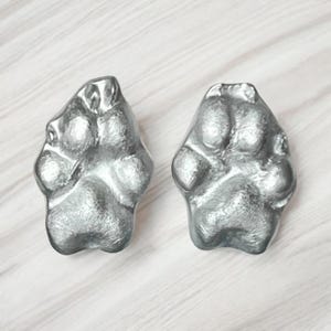 May include: Two silver-coloured paw print-shaped objects. The objects have a textured surface and are displayed on a light-coloured, wood-grain background. The paw prints are detailed and appear to be made of metal.