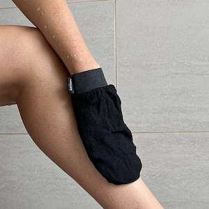 Puede incluir: Guante exfoliante negro con una banda elástica negra y la palabra "BIORASIA" impresa en la banda. El guante se está utilizando en la pierna de una persona.