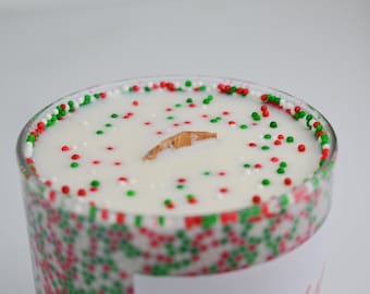 May include: A clear glass candle with a white wax interior, decorated with red, green, and white sprinkles around the rim and scattered on top. A wooden wick is centered in the wax. The candle is a festive item.