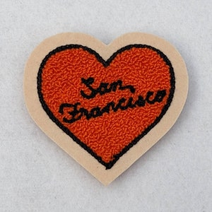 May include: A heart-shaped patch with a beige background and a black outline. The heart is filled with orange felt and has the words "San Francisco" embroidered in black.