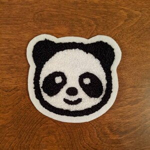 May include: A tufted panda bear face patch featuring a white center with black ears, eye patches, nose, and mouth. The design is outlined in a white border, creating a soft, textured appearance with a distinct contrast between the black and white yarn.
